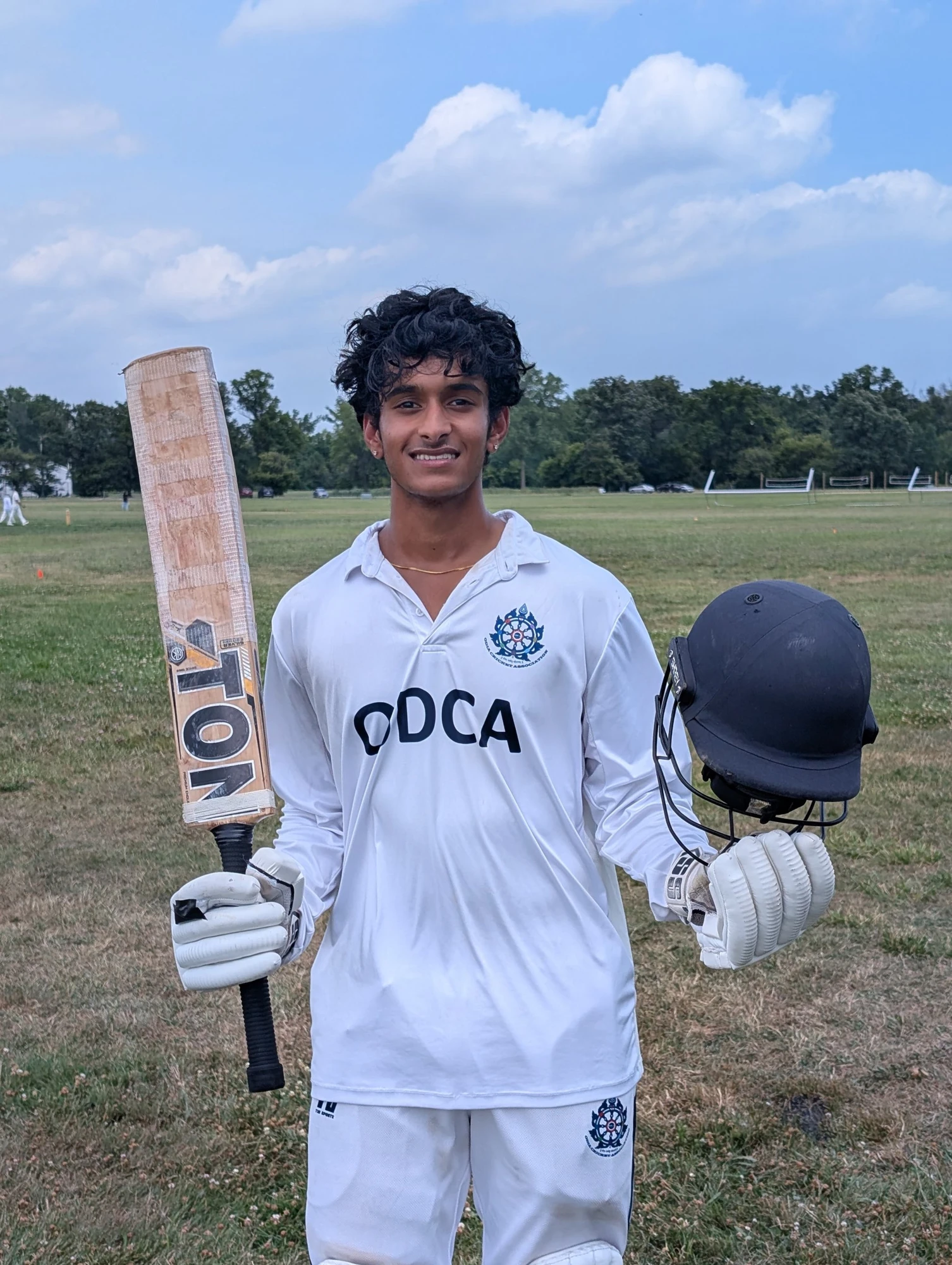 Dev in cricket whites holding his bat and helmet